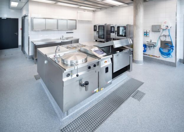 Eine große gewerbliche Küche mit modernen Edelstahlgeräten und umfangreicher Ausstattung für die Lebensmittelzubereitung auf beschichtetem Boden.