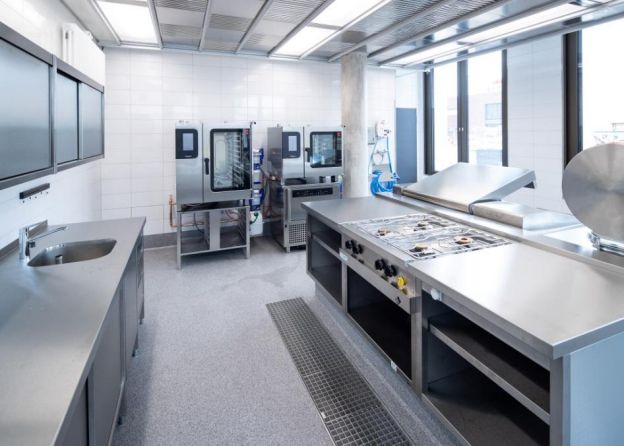 Eine große gewerbliche Küche mit modernen Edelstahlgeräten und umfangreicher Ausstattung für die Lebensmittelzubereitung auf beschichtetem Boden, von einer anderen Perspektive.